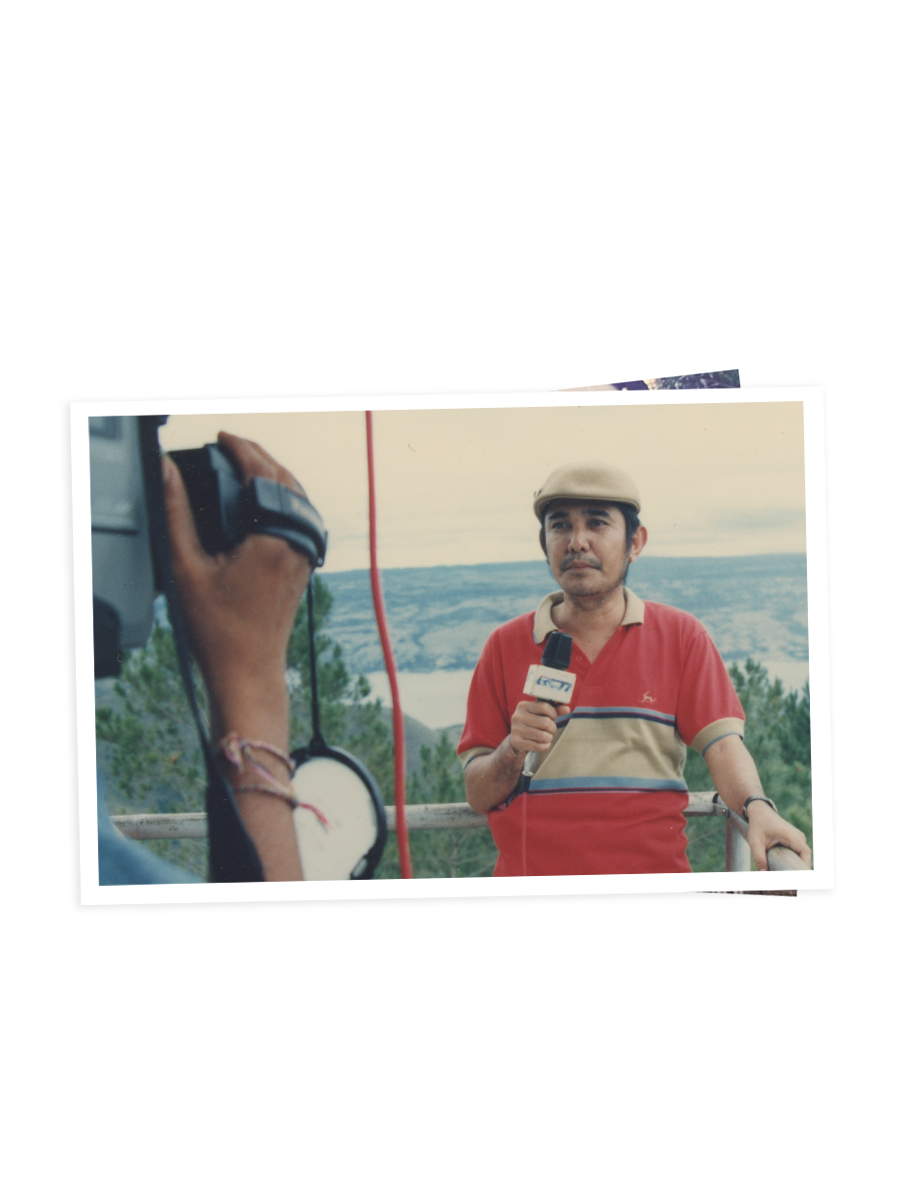 Ersa Siregar, wearing a cap and holding a microphone, reports outdoors with hills behind him.
